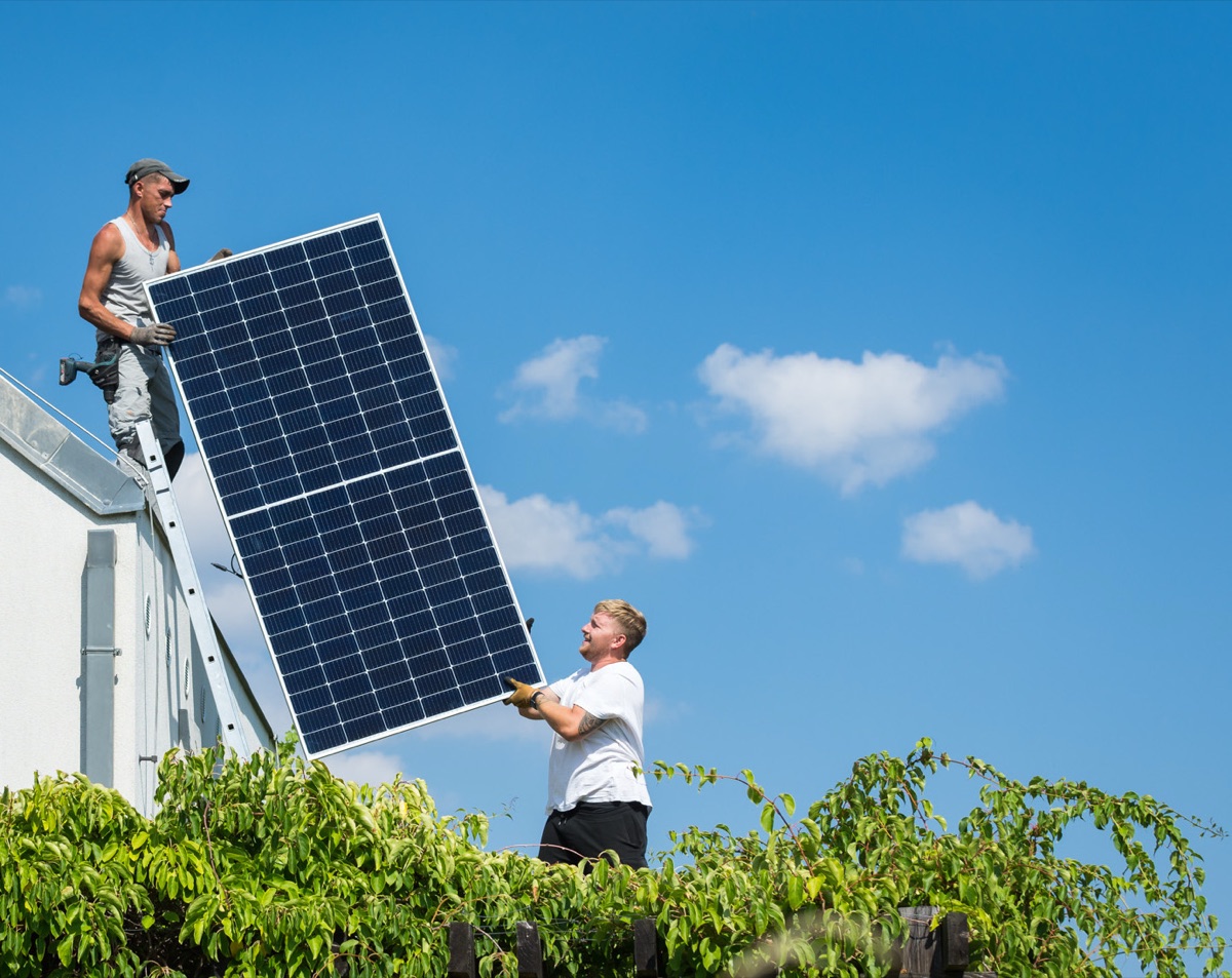 Dva montéři zvedají fotovoltaický panel na střechu.